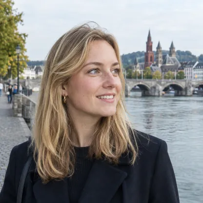 Portret van een lachende vrouw met halflang blond haar, met de Maas en een stenen brug op de achtergrond