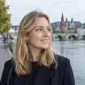 Portret van een lachende vrouw met halflang blond haar, met de Maas en een stenen brug op de achtergrond
